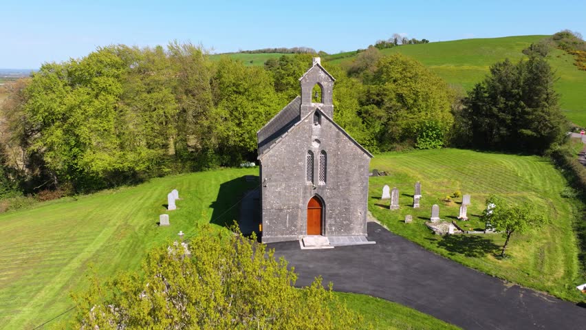 A historic stone church stands amidst lush green fields and ancient gravestones in County Laois, Ireland. Ideal for historical documentaries or cultural tourism content.