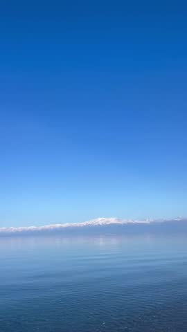 A breathtaking and peaceful landscape featuring a distant range of snow-covered mountains across the calm, clear blue waters of a vast lake.