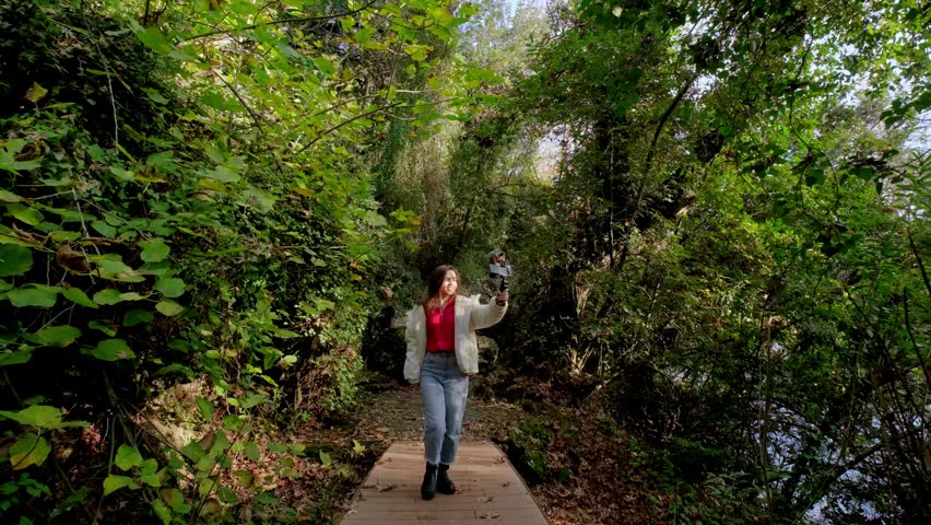Female vlogger stands on a forest path beside a green pond and waterfall, holding a vlog camera while recording outdoor travel and nature content among mossy rocks and lush trees in daylight.