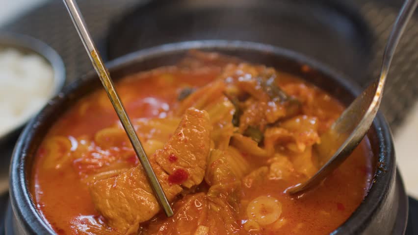 Metal chopsticks and a spoon lift pork and kimchi from a steaming hot stone bowl
