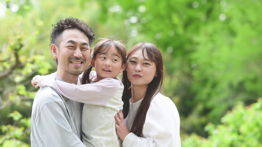 A Japanese family of three smiling and standing close together against a backdrop of beautiful fresh greenery. A family image capturing the happiness and bond between father, mother, and daughter. A close-up shot with the family looking directly at the camera.