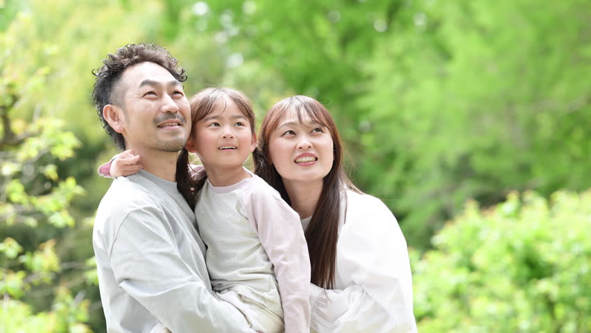 A Japanese family of three smiling and standing close together against a backdrop of beautiful fresh greenery. A family image capturing the happiness and bond between father, mother, and daughter. A close-up shot with the family looking directly at the camera. A slow-motion shot of them looking up and gazing toward the future.
