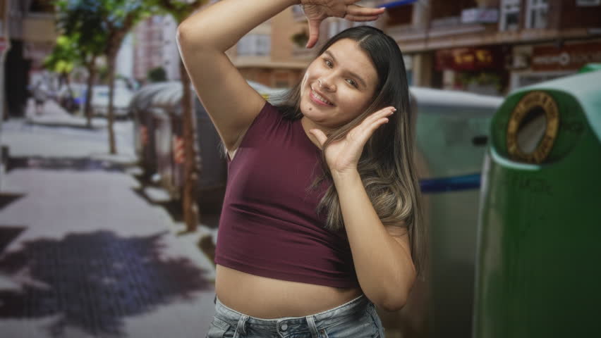 Young woman framing her face with both hands, maroon crop top exposing stomach on a busy street near a green recycling bin and storefronts; playful confidence.