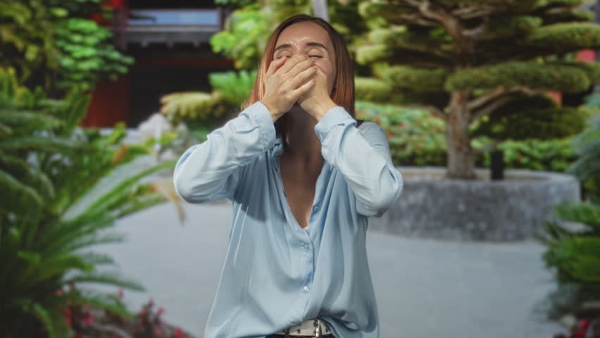 Woman covers mouth with both hands and wide eyes in a building courtyard with trimmed bonsai planter, wearing blue blouse and belt; surprise awe.