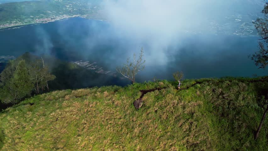 Lone hiker walking along narrow mountain ridge trail on steep grassy slope with crater lake below and cloud drifting across the ridge in high altitude landscape, aerial arc drone footage