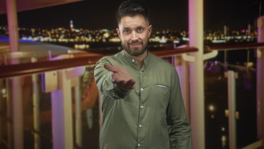 Man with outstretched hand offering palm on building balcony at night, smiling in a green shirt and beard; invitation welcome warmth.