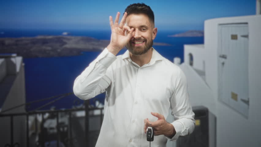Hispanic man holding car keys and making ok sign on white building terrace overlooking blue sea and island; confidence travel vacation.