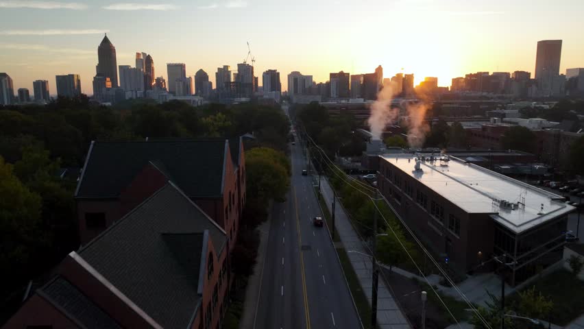 Aerial view of a vibrant city street with moving vehicles during sunrise. The Atlanta skyline looms in background, steam rises from a building, and golden light bathes scene, showcasing dynamic urban life.