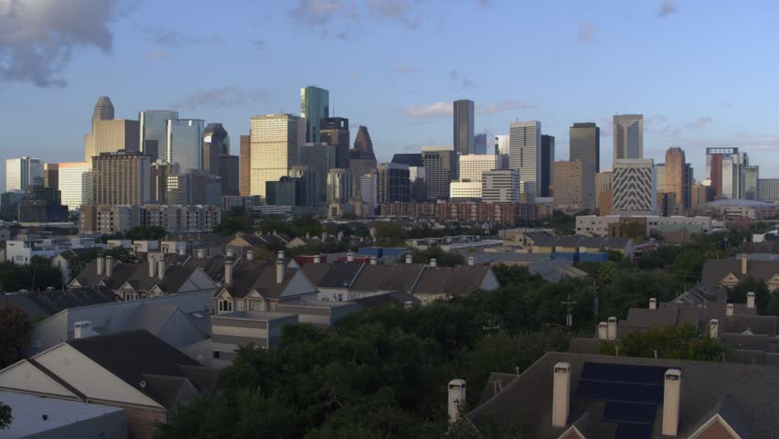 Static cinematic shot of Houston skyline at dusk. Urban cityscape features towering skyscrapers including Heritage Plaza and Enterprise Plaza. Residential houses sit in foreground of city.
