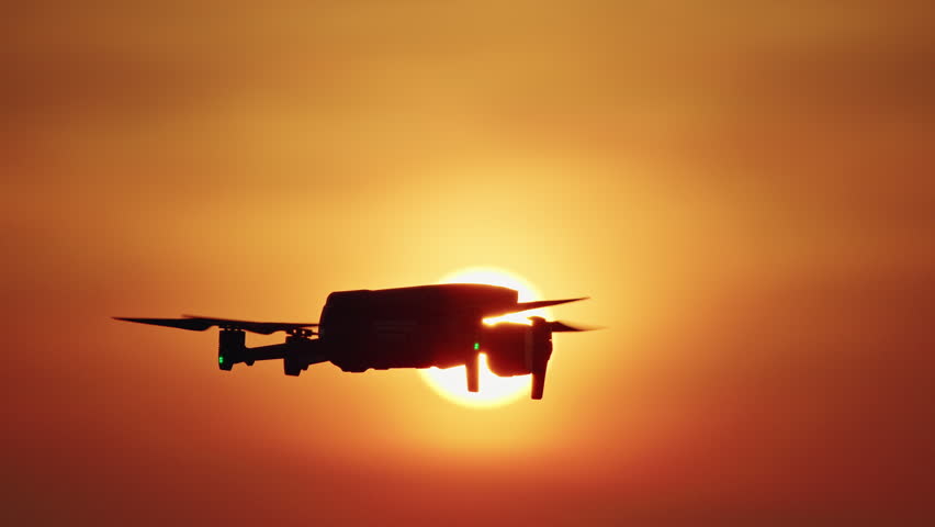 Drone silhouette flying against bright sun at sunset with orange sky.