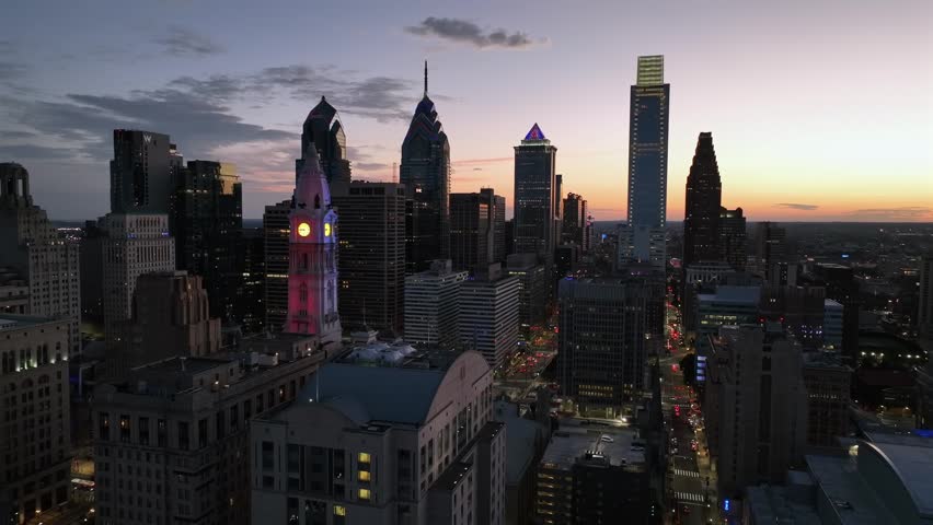 Dynamic aerial view of Philadelphia cityscape at twilight. Illuminated city hall tower glows with vibrant colors against urban skyscrapers. Evening city lights create atmosphere.