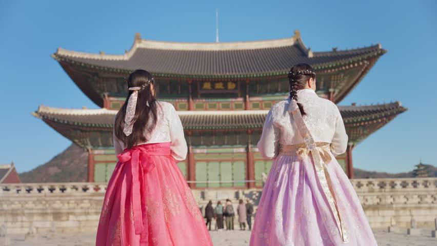 Two women in elegant traditional Korean hanbok dresses with pink and lavender skirts admire the majestic Gyeongbokgung Palace under a clear blue sky in Seoul, tourists visible in the distance.