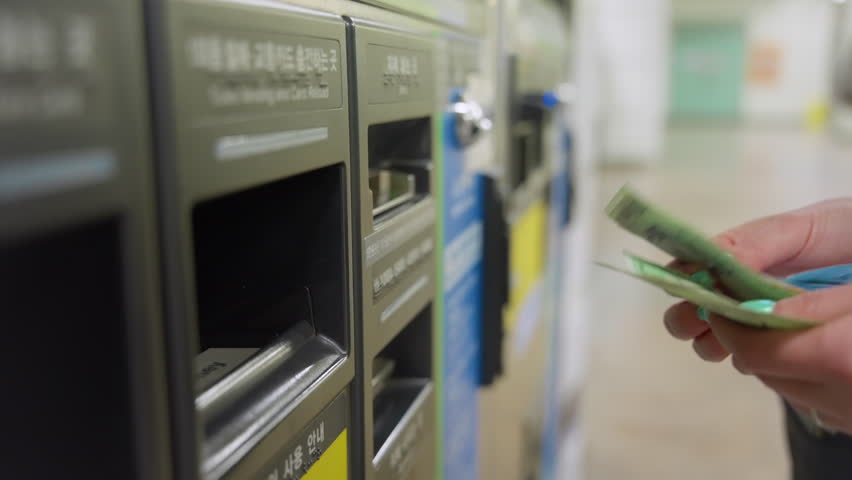 Woman with green manicured nails inserts Korean won banknotes into subway ticket machine in Seoul South Korea. Metro station interior with travelers in background and modern automated kiosk system.