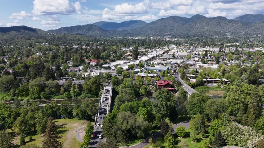 Downtown Grants Pass city skyline with Caveman Bridge over Rogue River with lush green trees and Southern Oregon mountains in background. Wide aerial drone wide shot.