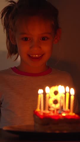 Portrait of little happy girl blows out burning candles on cake on birthday and laughs. Childs Birthday celebration. Vertical shot