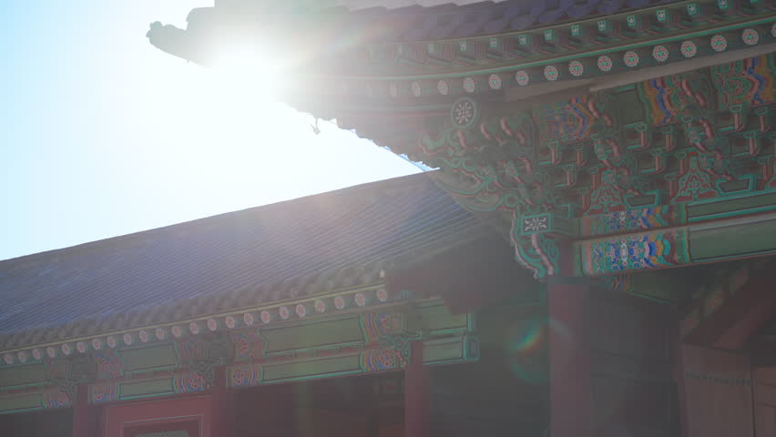 Sunlight illuminates intricate painted eaves and traditional roof tiles at Gyeongbokgung Palace in Seoul, South Korea. Guardian figurines stand along the ridge with vibrant dancheong patterns visible.
