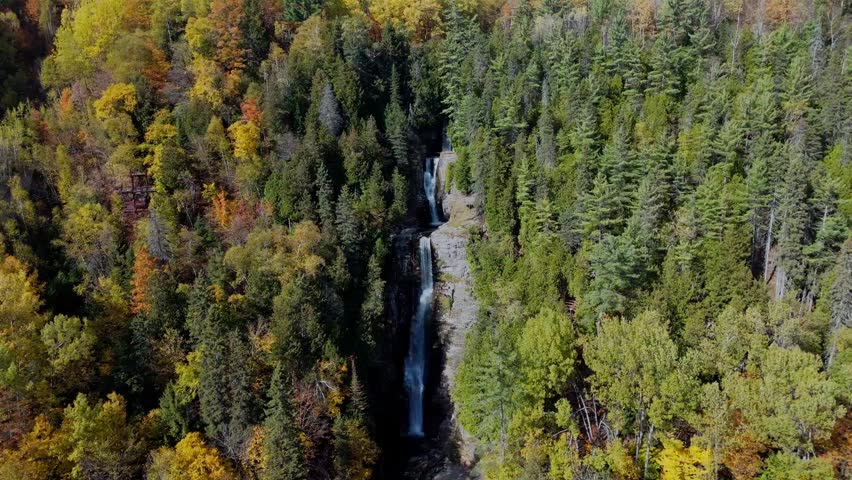 Drone flying backwards and upwards from the waterfall to reveal the vast autumn forest, Mont-Sainte-Anne ski resort area and scenic Quebec mountains.