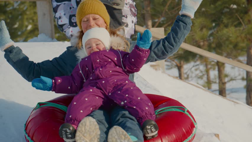 Mother and child laugh while sledding down snowy hill. Happy mother holds child in snow-filled sled. Joyful toddler waves arms as they slide. Perfect for winter family, holiday