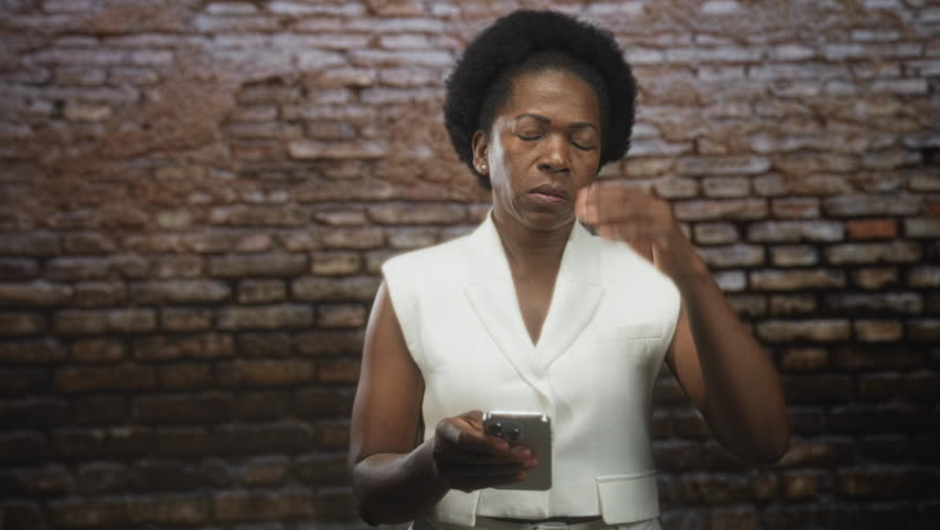 Woman holding smartphone with both hands and typing with thumbs while standing against brick wall in studio wearing white sleeveless blazer; quiet concentration.