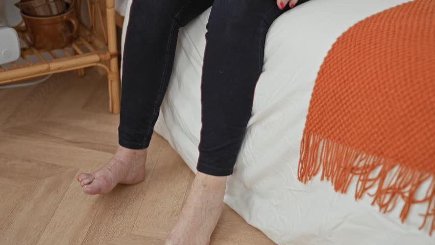 Woman sitting on bed, leaning forward and holding her knee with both hands while looking down in a bedroom with pillows and books; concern aging.