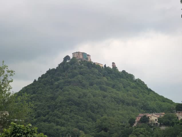 A hill covered in vegetation, topped by an imposing historic stone structure. A charming and scenic atmosphere, with small buildings nestled in the greenery at the foot of the promontory.