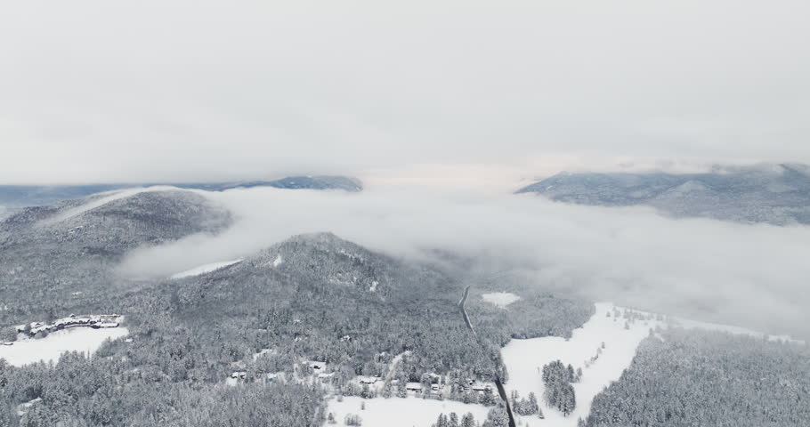 Drone fly in over a snow-covered Adirondack valley with scattered homes and forest as low winter clouds roll across mountain ridges near Lake Placid, New York.