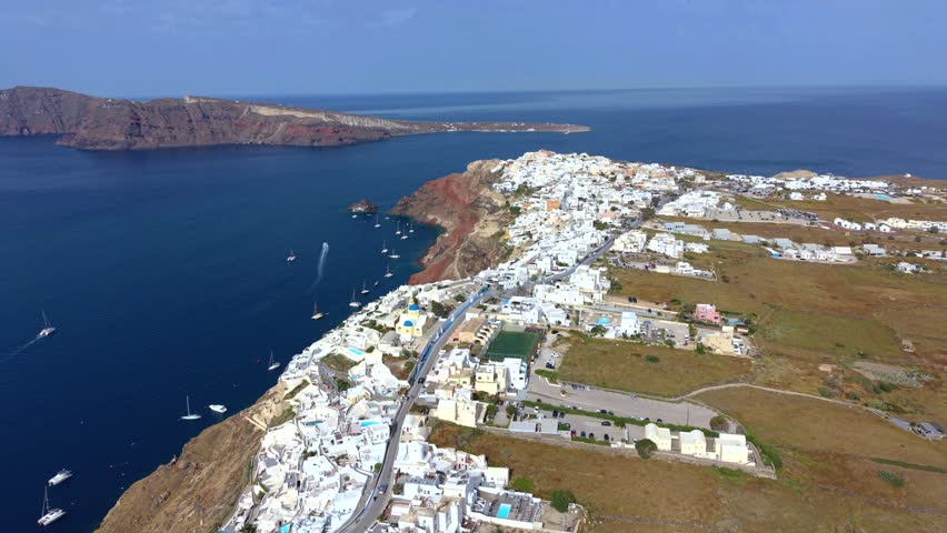 Aerial drone view of Santorini island near Oia, Greece, during a sunny day in May. Authentic natural landscape featuring traditional white houses, coastal cliffs, deep blue Aegean Sea, surrounding islands, and less touristy residential and desert areas near the famous hiking trail between Oia and Fira. Real unaltered footage capturing the genuine atmosphere and geography of Santorini.