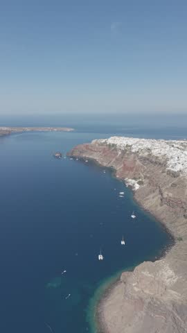 Aerial drone view of Santorini island near Oia, Greece, during a sunny day in May. Authentic natural landscape featuring traditional white houses, coastal cliffs, deep blue Aegean Sea, surrounding islands, and less touristy residential and desert areas near the famous hiking trail between Oia and Fira. Real unaltered footage capturing the genuine atmosphere and geography of Santorini.