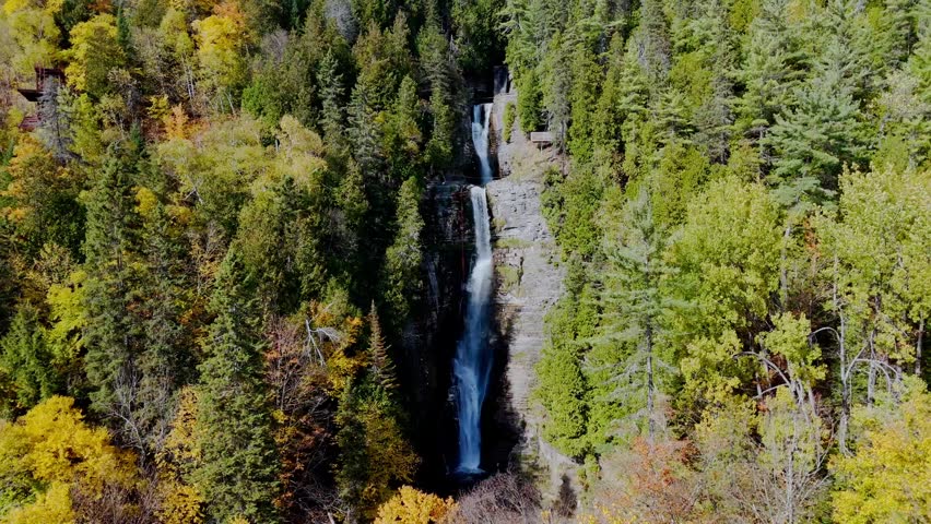 Drone flying backwards and upwards from the Jean-Larose waterfall to reveal the stunning autumn forest and rocky gorge in Quebec, Canada.
