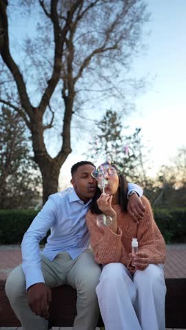 Young diverse couple sitting on a park bench and blowing soap bubbles. They are laughing and having a good time together during a date