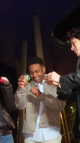 Joyful multiethnic friends toasting and drinking colorful alcohol shots. Young people having fun at a party on a city street at night