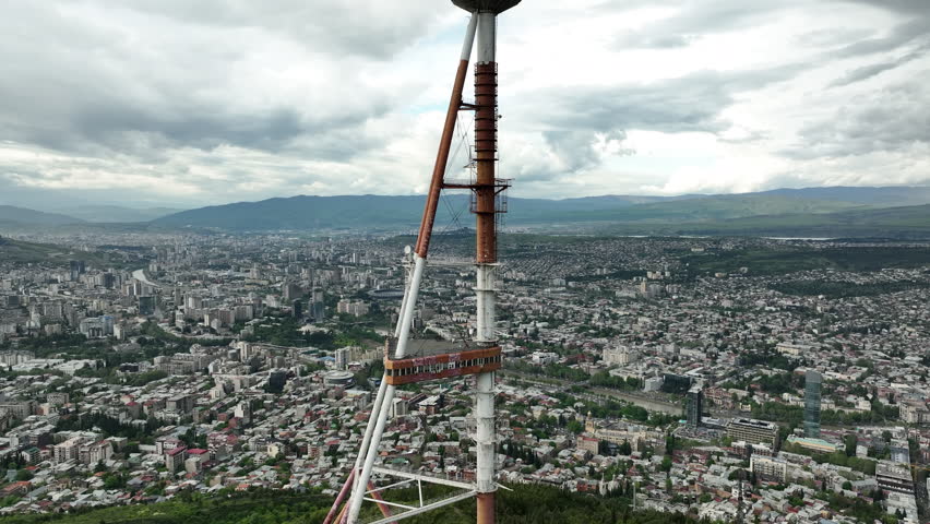Aerial shot of the iconic Tbilisi TV Broadcasting Tower on Mtatsminda Mountain overlooking the city architecture during daytime.
