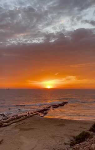 HDR footage of sunset over Atlantic Ocean with golden light reflecting on calm sea waves. Rocky beach shoreline with silhouette of person walking. High dynamic range captures rich colors and details. Perfect for travel, tourism, and nature video projects.