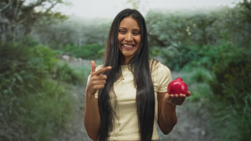 Woman holding a pomegranate and pointing finger toward viewer on a forest path, smiling while offering the fruit; playful invitation.