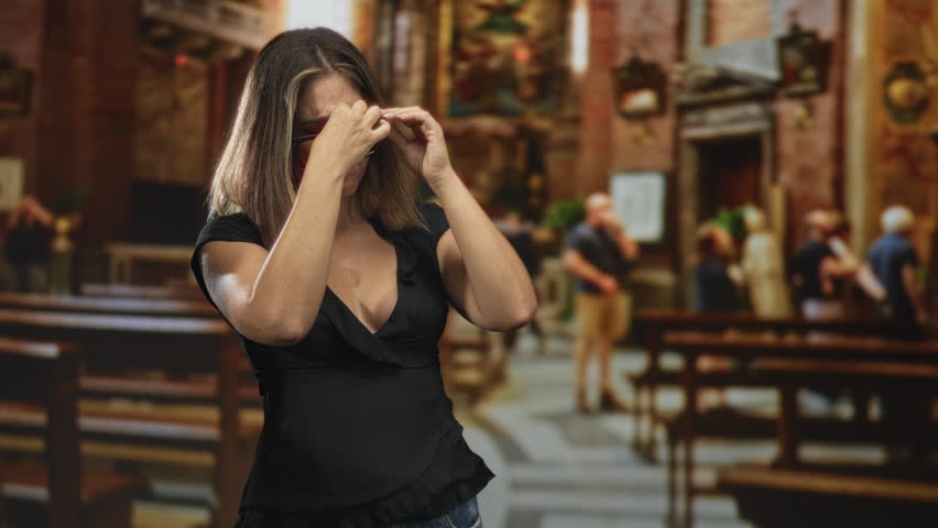 Woman rubbing forehead with hand while adjusting glasses inside ornate church building, black blouse visible; fatigue.