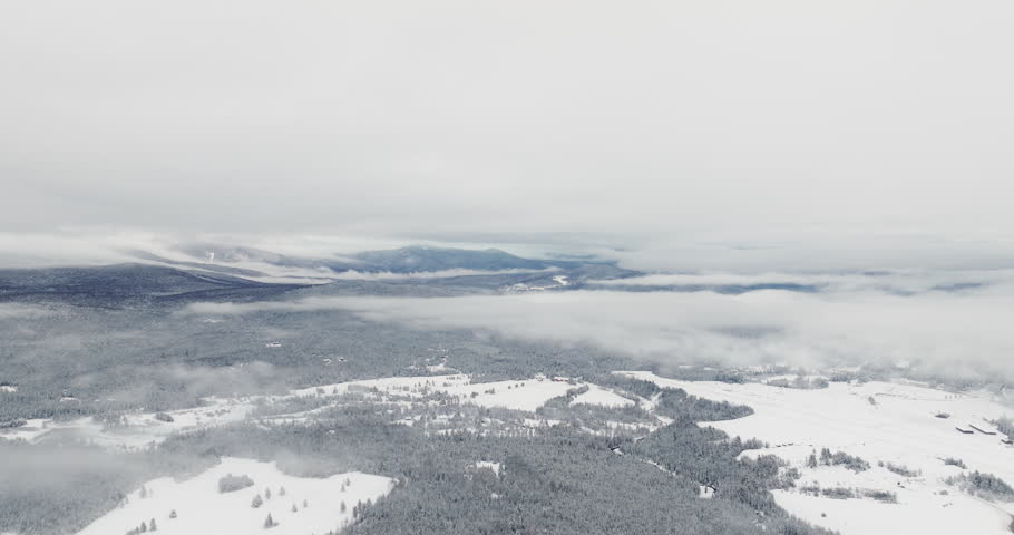 Drone flies over snow-covered farmland and forest with distant Adirondack mountains partially hidden beneath layered winter clouds after fresh snowfall near Lake Placid, New York.