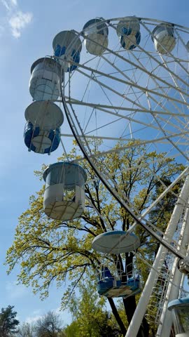 White ferris wheel with blue cabins rotating against blue sky and green trees in city amusement park during sunny spring day