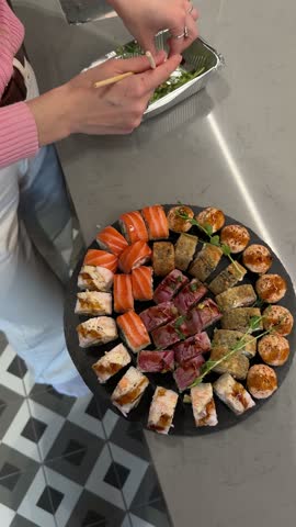 4K close up video of a young woman chef hand decorating a large mixed sushi set platter with green microgreens sprouts on a modern kitchen counter table