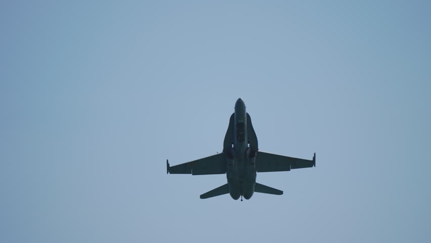 Military fighter jet flies directly overhead against clear blue sky. Camera follows aircraft as it gets closer, revealing details of its undercarriage and wings. Powerful and dynamic shot