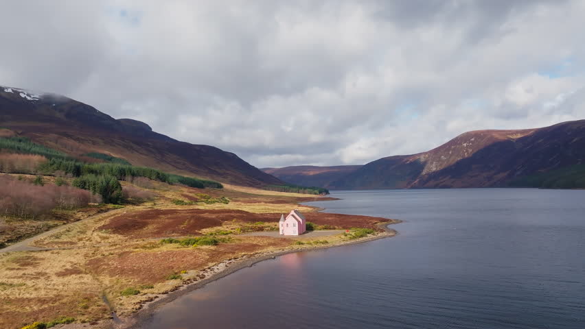 Drone flyover of the Pink House at Loch Glass, Scottish Highlands, partly cloudy sky casting a mix of light and shadow across the lochside scene