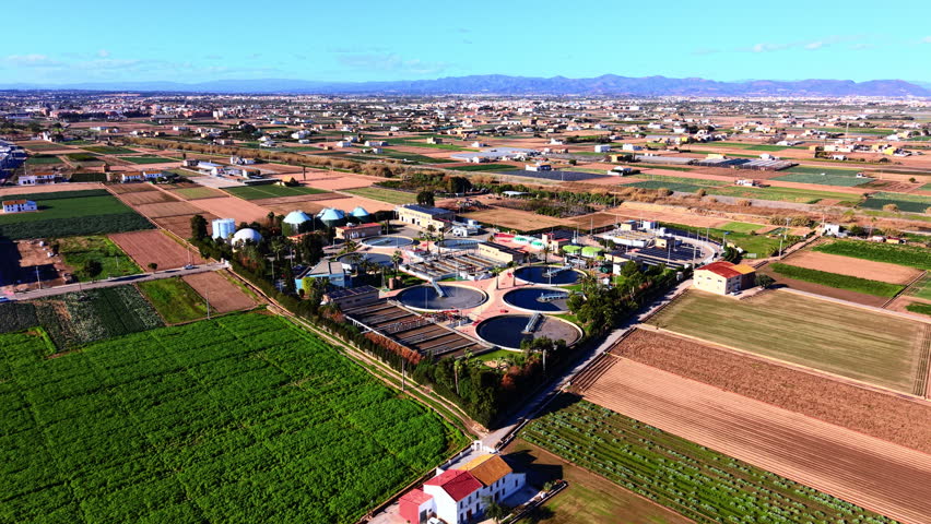 Modern sewage processing plant surrounded by green cultivated farm fields under a clear blue sky. Industrial water treatment facility located in agricultural landscape.