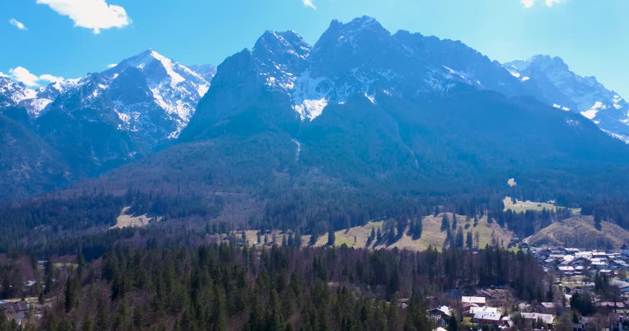 Mountains tower over a valley filled with trees and a small village. Snow caps the peaks. The weather is clear with bright sunlight illuminating the landscape. The Alps, Germany.