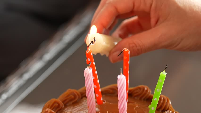 Hands using a lit white candle to ignite colorful birthday candles on a caramel birthday cake. Warm indoor celebration scene with soft light, festive atmosphere and focus on birthday tradition