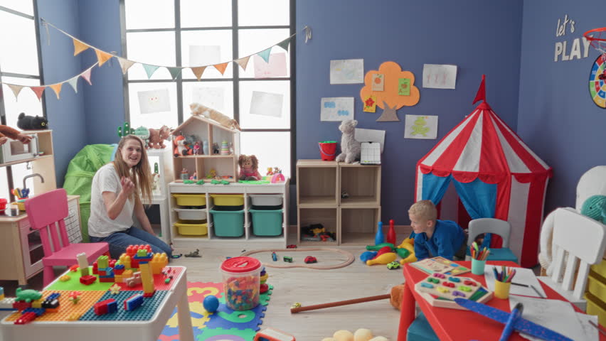 Woman kneeling and reaching toward boy crawling among scattered blocks, toy train and play tent in preschool playroom; nurturing play.