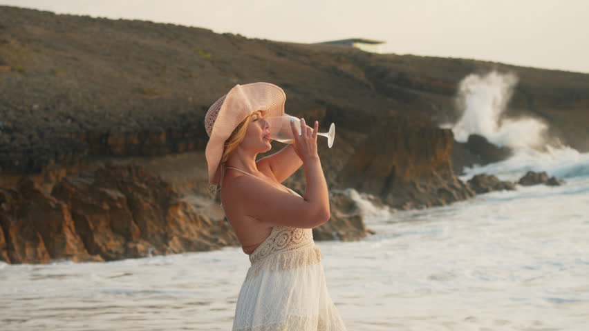 Elegant woman in white dress standing on sandy beach near rocky cliffs during sunset. Side view with warm golden light, holding a glass of wine and enjoying calm ocean atmosphere.