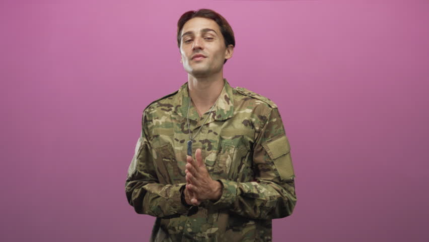 Soldier man in camouflage jacket clapping hands in a pink studio with dog tags visible and relaxed expression; pride duty.