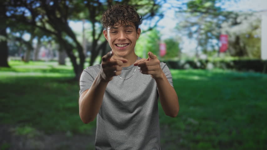 Teen boy points finger at camera while smiling, wearing gray tshirt and necklace in front of a building on a sunny lawn; playful confidence.