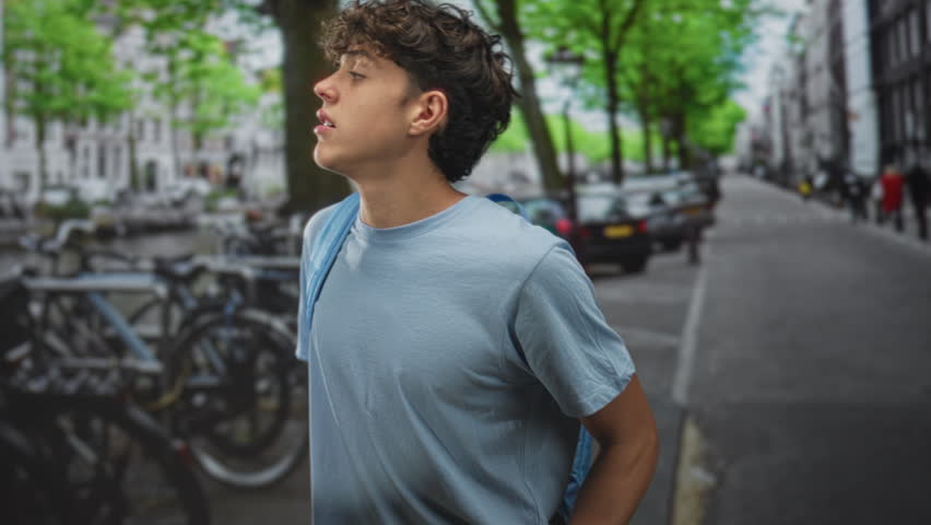 Teenage man in blue shirt with backpack looking sideways, head tilted and eyes half closed near parked bicycles and cars on street; contemplation.