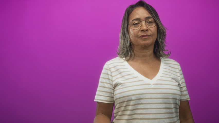Hispanic middle age woman points finger to chest, wearing striped shirt and glasses against a magenta studio backdrop and soft lighting; confidence affirmation.