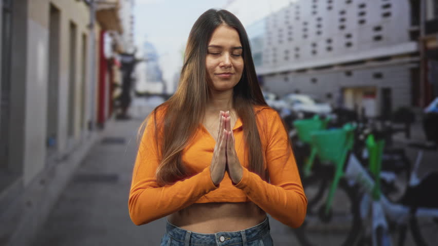 Woman with hands pressed together in prayer on a street, orange cropped top and jeans, eyes closed in a quiet moment; serenity gratitude.